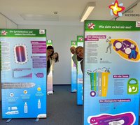 Die genannten Personen stehen zwischen großen Ausstellungswänden und schauen in die Kamera.