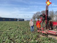 Zwei Männer stehen auf einem freien, grün bewachsenen Feld neben einer roten Maschine, die Erdbohrungen vornehmen kann. Im Hintergrund eine eine große Halle mit der Aufschrift "Rietberger Möbelwerke" zu erkennen.