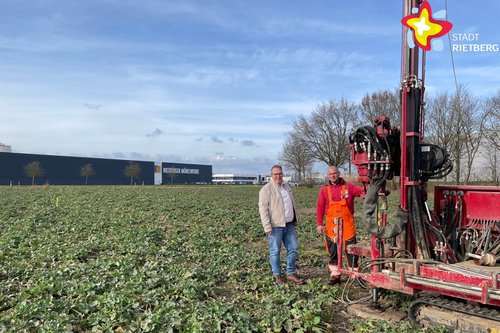 Zwei Männer stehen auf einem freien, grün bewachsenen Feld neben einer roten Maschine, die Erdbohrungen vornehmen kann. Im Hintergrund eine eine große Halle mit der Aufschrift "Rietberger Möbelwerke" zu erkennen.