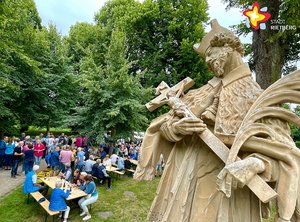 Die Statue des St. Johannes Nepomuk im Vordergrund scheint auf die Besucher zu blicken, die im Hintergrund unter Bäumen an Tischen sitzen.