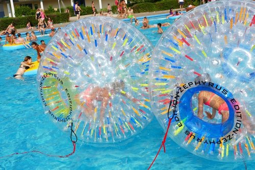 Große Bälle, in denen Kinder sich bewegen, treiben auf dem Wasser eines Freibadbeckens. im Hintergrund spielen und toben weitere Kinder auf Schwimmreifen.