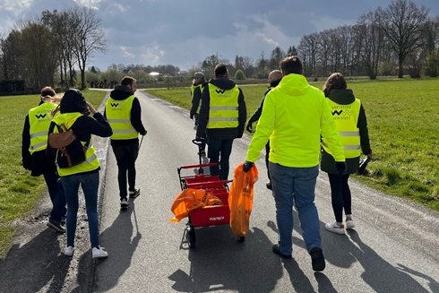 Eine Gruppe von acht Personen, alle in dunklen Jacken mit gelben Westen bekleidet, läuft eine Straße zwischen grünen Wiesen entlang, die Rücken zur Kamera. Sie ziehen in ihrer Mitte einen Bollerwagen.
