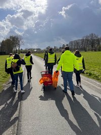 Eine Gruppe von acht Personen, alle in dunklen Jacken mit gelben Westen bekleidet, läuft eine Straße zwischen grünen Wiesen entlang, die Rücken zur Kamera. Sie ziehen in ihrer Mitte einen Bollerwagen.