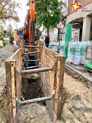 Zwischen zwei stabilen Schutzwänden ist eine Rohrgrube inmitten einer Straße zu erkennen. im Hintergrund steht einBagger.