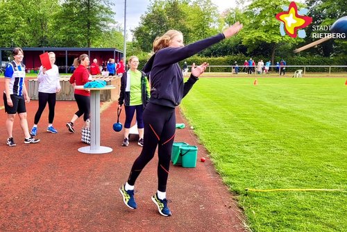 Mehrere sportliche gekleidete Menschen sehen auf einem Sportplatz zu, wie eine junge Frau einen Schleuderball wirft.