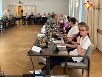 Im Ratssaal sitzen Schülerinnen und Schüler gemeinsam mit Politikern an Tischen und halten eine Ratssitzung ab. In dem großzügig dimensionierten Raum liegt Parkett. Die Wände sind weiß gestrichen.