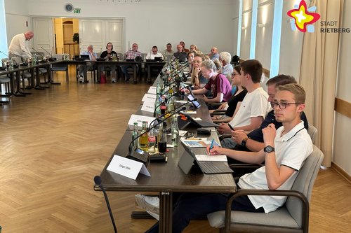 Im Ratssaal sitzen Schülerinnen und Schüler gemeinsam mit Politikern an Tischen und halten eine Ratssitzung ab. In dem großzügig dimensionierten Raum liegt Parkett. Die Wände sind weiß gestrichen.