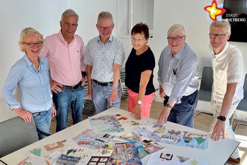 Sechs Frauen und Männer stehen rund um einen Tisch, der bedeckt ist mit gemalten Bildern.