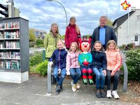 Lia, Jule, Ida, Pia und Marlene sitzen unter freiem Himmel auf einer Bank - die Rieti-Figur »Westi« in der Mitte. Kunstwerkstatt-Leiterin Karin Wolf und Wilfried Dörhoff von der Stadt Rietberg stehen links und rechts daneben und blicken ebenfalls in die Kamera.