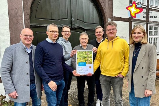 Sechs Männer und eine Frau stehen nebeneinander vor einem Fachwerkhaus und halten ein kleines Plakat in der Mitte.