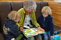 Eine Dame sitzt auf einem Sofa, ein aufgeschlagenes Bilderbuch auf dem Schoss. Rechts und links von ihr sitzen zwei Kinder, die mit ihr zusammen ins Buch schauen.
