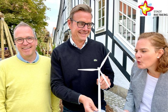 Rüdiger Ropinski, Abteilungsleiter Stadtentwicklung, und Bürgermeister Andreas Sunder stehen vor dem Historischen Rathaus. Sunder hält das Modell eines Windrades in der Hand, gegen welches Klimaschutzmanagerin Svenja Schröder von der Seite pustet.