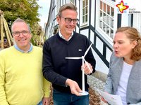 Rüdiger Ropinski, Abteilungsleiter Stadtentwicklung, und Bürgermeister Andreas Sunder stehen vor dem Historischen Rathaus. Sunder hält das Modell eines Windrades in der Hand, gegen welches Klimaschutzmanagerin Svenja Schröder von der Seite pustet.