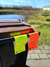 Ein gelber und ein roter Zettel hängen nebeneinander an den Griffen einer Mülltonne.