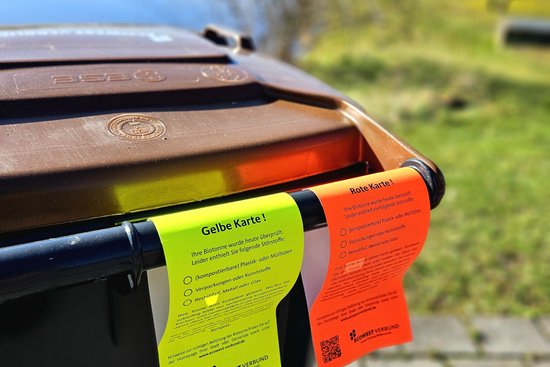 Ein gelber und ein roter Zettel hängen nebeneinander an den Griffen einer Mülltonne.