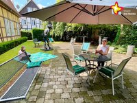 Eine Dame sitzt mit einem Buch in den Händen unter einem Sonnenschirm am Tisch, während links davon eine Frau auf einer grünen Picknickdecke sitzt.