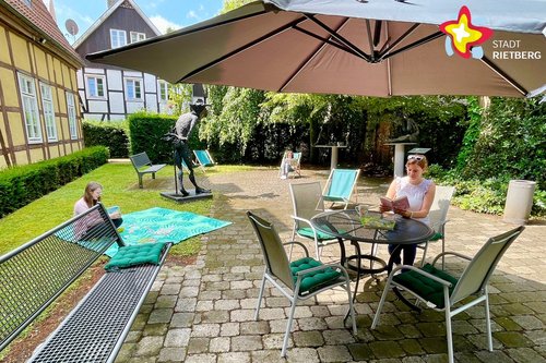 Eine Dame sitzt mit einem Buch in den Händen unter einem Sonnenschirm am Tisch, während links davon eine Frau auf einer grünen Picknickdecke sitzt.