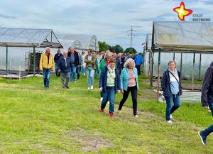 Frauen und Männer spazieren über eine grüne Wiese, im Hintergrund sind Gewächshäuser zu sehen.