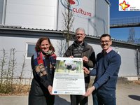 Klimaschutzmanagerin Svenja Schröder, Michael Kaune aus der Abteilung Hochbau und Gartenschaupark-Geschäftsführer Johannes Wiethoff stehen mit einem Plakat in der Hand vor der Cultura, im Hintergrund ist ein neu gepflanzter Baum zu sehen.