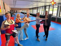 Die genannten Personen stehen in Selbstverteidigungspose im Trainingsraum. Im Hintergrund ist ein großer Wandspiegel zu sehen. Der Boden ist mit blau-roten Matten ausgelegt.