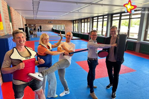 Die genannten Personen stehen in Selbstverteidigungspose im Trainingsraum. Im Hintergrund ist ein großer Wandspiegel zu sehen. Der Boden ist mit blau-roten Matten ausgelegt.