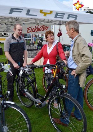 Unter einem Pavillon lässt sich ein Ehepaar mit einem Fahrrad in der Hand von einem Fahrradhändler beraten. Im Hintergrund hängt ein Plakat mit der Aufschrift "Eickhölter".