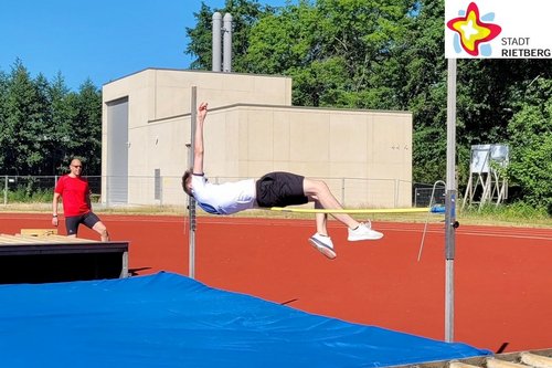 Bei strahlendem Sonnenschein springt ein jugendlicher Sportler auf einer Outdoor-Sportanlage rückwärs über eine Hochsprung-Stange.