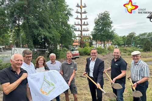 Die genannten Personen halten entweder einen Spaten mit Erde in der Hand oder (die anderen) einen Lageplan. Alle schauen in die Kamera. Im Hintergrund ist der Handwerkerbaum zu sehen.