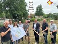 Die genannten Personen halten entweder einen Spaten mit Erde in der Hand oder (die anderen) einen Lageplan. Alle schauen in die Kamera. Im Hintergrund ist der Handwerkerbaum zu sehen.
