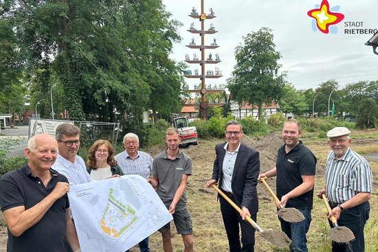 Die genannten Personen halten entweder einen Spaten mit Erde in der Hand oder (die anderen) einen Lageplan. Alle schauen in die Kamera. Im Hintergrund ist der Handwerkerbaum zu sehen.