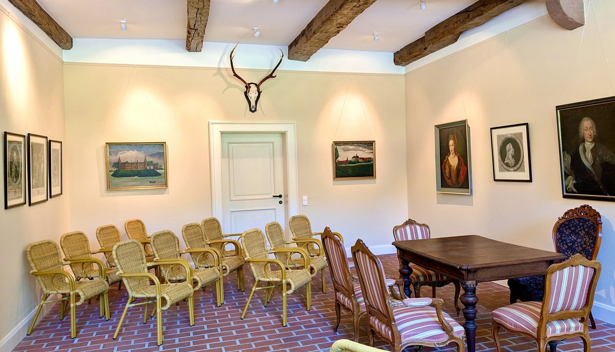 Leere Korbstühle sind in Reihen aufgestellt. Ein dunkler Tisch mit vier antiken Stühlen drumherum steht am Rand. An der Wand hängen altertümliche Bilder.