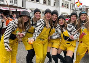 Eine Gruppe Frauen in gestreiften Pullis und gelben Latzhosen und mit Mützen auf dem Kopf lacht in die Kamera.