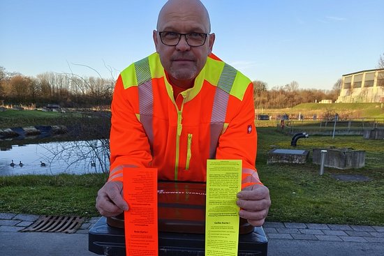 Ein Mann in orangefarbener Jacke lehnt auf einer schwarzen Mülltonne und hält je eine rote und gelbe in den Händen.