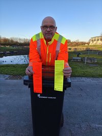 Ein Mann in orangefarbener Jacke lehnt auf einer schwarzen Mülltonne und hält je eine rote und gelbe in den Händen.
