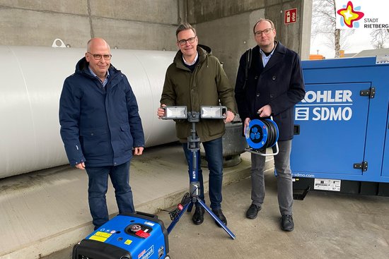 Drei Männer stehen vor einem großen blauen Notstromaggregat und schauen frontal in die Kamera. Zu sehen sind weitere Utensilien wie eine Kabeltrommel, Heizlüfter und Lampen. Oben rechts auf dem Foto befindet sich das Logo der Stadt Rietberg