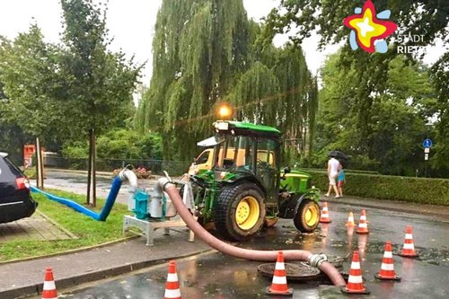 Ein grüner Traktor steht am Straßenrand. Rot-weiß gestreifte Pilonen stehen um einen geöffneten Gullideckel herum.