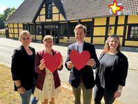 Die genannten Personen stehen vor einem Fachwerkgebäude auf dem Rasen und schauen in die Kamera. Zwei Personen halten rote Herzen in die Kamera.