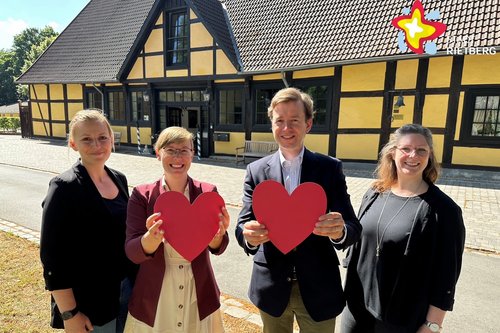 Die genannten Personen stehen vor einem Fachwerkgebäude auf dem Rasen und schauen in die Kamera. Zwei Personen halten rote Herzen in die Kamera.