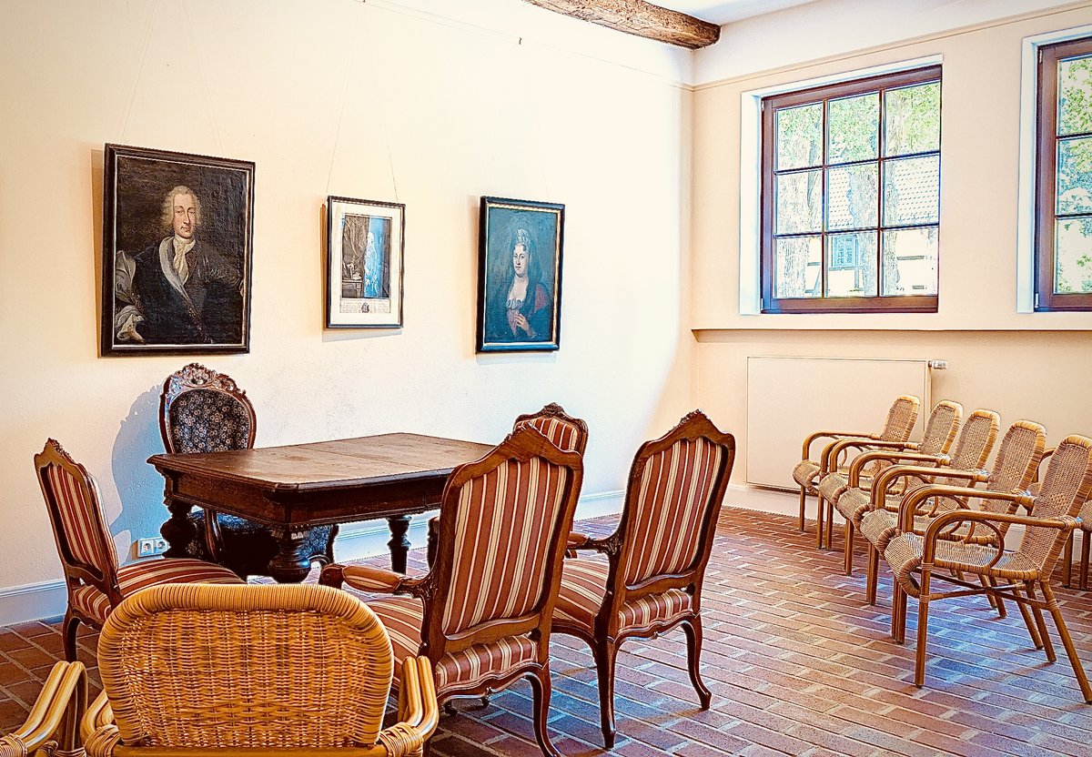 Ein dunkler Tisch mit vier antiken Stühlen drumherum steht am Rand eines Zimmers. An der Wand hängen altertümliche Bilder. Eine Reihe mit leeren Korbstühlen ist aufgestellt.