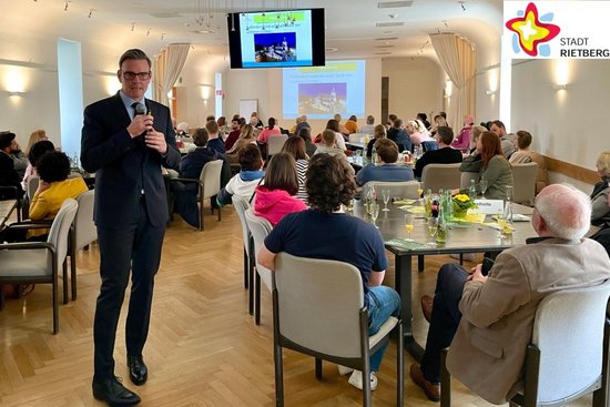 Bürgermeister Andreas Sunder steht mit einem Mikrofon in der Hand mitten im Ratssaal des Prygymnasiums. Um ihn herum sind Tischgruppen drapiert, besetzt mit Menschen unterschiedlichen Alters, die seinem Vortrag zuhören.