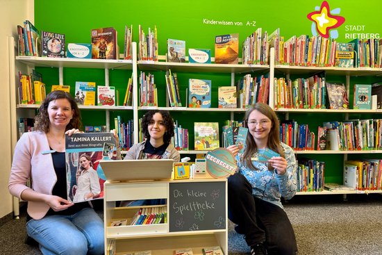 Die drei Bibliotheksmitarbeiterinnen knien vor einem Bücherregal, in der Hand haben sie Bibliotheksausweise, Lesezeichen und ein Plakat zur Tamina-Kallert-Lesung. Zu sehen ist auch ein Holzregal mit der Aufschrift "Die kleine Spieltheke", an der Kinder Bücherei spielen können.