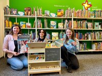 Die drei Bibliotheksmitarbeiterinnen knien vor einem Bücherregal, in der Hand haben sie Bibliotheksausweise, Lesezeichen und ein Plakat zur Tamina-Kallert-Lesung. Zu sehen ist auch ein Holzregal mit der Aufschrift "Die kleine Spieltheke", an der Kinder Bücherei spielen können.