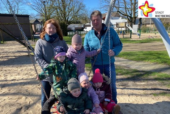 Zwei Erwachsene stehen hinter der neuen Nestschaukel, fünf Kinder sitzen in der Nestschaukel. Im Hintergrund ist blauer Himmel zu sehen, unter der Schaukel heller Sand. Die Sonne scheint.