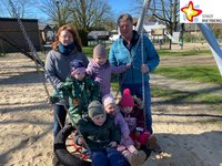 Zwei Erwachsene stehen hinter der neuen Nestschaukel, fünf Kinder sitzen in der Nestschaukel. Im Hintergrund ist blauer Himmel zu sehen, unter der Schaukel heller Sand. Die Sonne scheint.