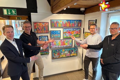 Die genannten Personen zeigen auf bunte Bilder von James Rizzi, die an einer Wand im Museum hängen. Sie schauen in die Kamera.