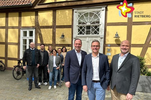 Neun Frauen und Männer stehen nebeneinander vor einer gelben Fachwerkfassade.