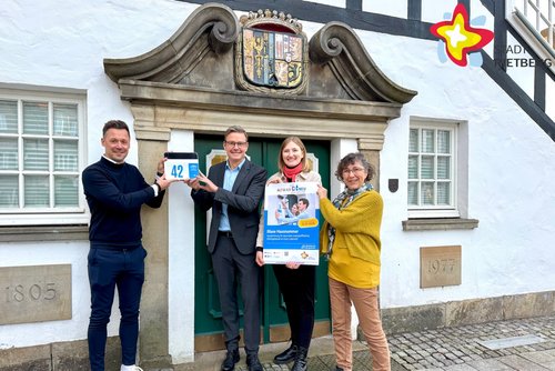 Mario Aschhoff und Bürgermeister Andreas Sunder halten symbolisch eine Blaue Hausnummer an das Historische Rietberger Rathaus. Rietbergs Klimaschutzmanagerin Wibke Kordtomeikel und Michaela Prelle von der Energieberatung der Verbraucherzentrale NRW stehen daneben und halten ein Werbeplakat in den Händen.