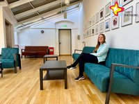 Julia Schmitfranz sitzt auf einem grünen Sofa, über dem mehrere Bilderrahmen an einer weißen Wand hängen. Vor ihr steht ein niedriger Tisch.