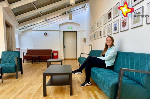 Julia Schmitfranz sitzt auf einem grünen Sofa, über dem mehrere Bilderrahmen an einer weißen Wand hängen. Vor ihr steht ein niedriger Tisch.