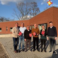 Die genannten Personen stehen vor dem Neubau und schauen in die Kamera. Der Neubau ist rot geklinkert, die Sonne schein.
